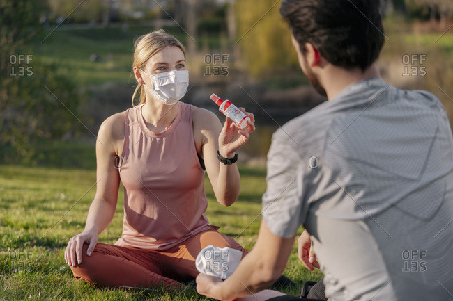 Woman wearing face mask showing hand sanitizer to man while sitting at park