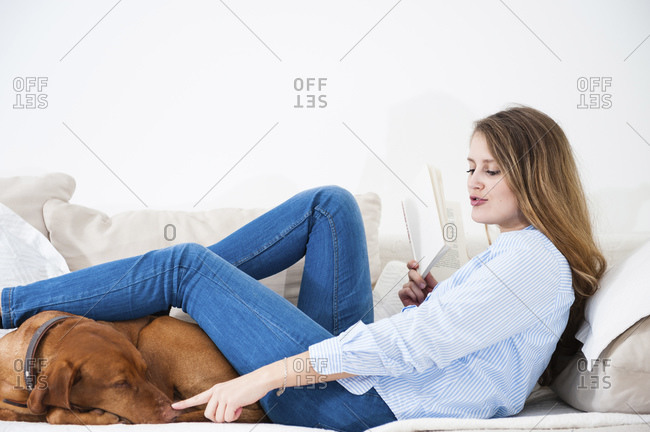 Playful young woman touching dog's snout while sitting with book on sofa at home