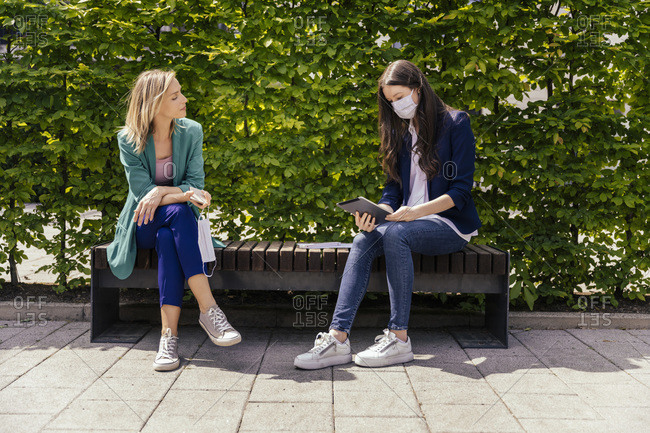 Two businesswomen sitting on bench outside and keeping their distance while wearing face mask