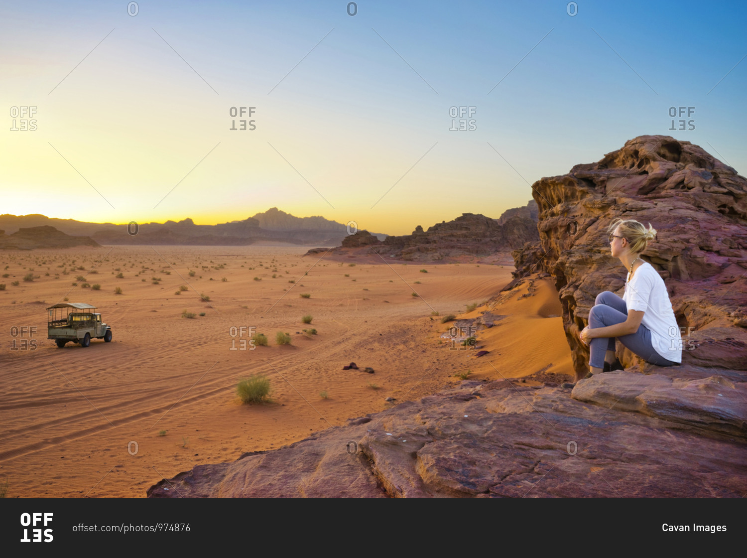 A woman sits on a rocky mountain top watching the sun set in the Wadi Rum desert, a truck is in the distance