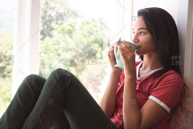 Mixed race woman spending time at home self isolating and social distancing in quarantine lockdown during coronavirus covid 19 epidemic, sitting on window seat drinking coffee in sitting room.