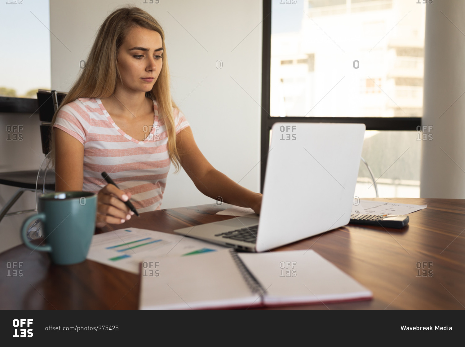 Caucasian woman sitting by a table, using a laptop and writing on a sheet of paper. Social distancing and self isolation in quarantine lockdown.