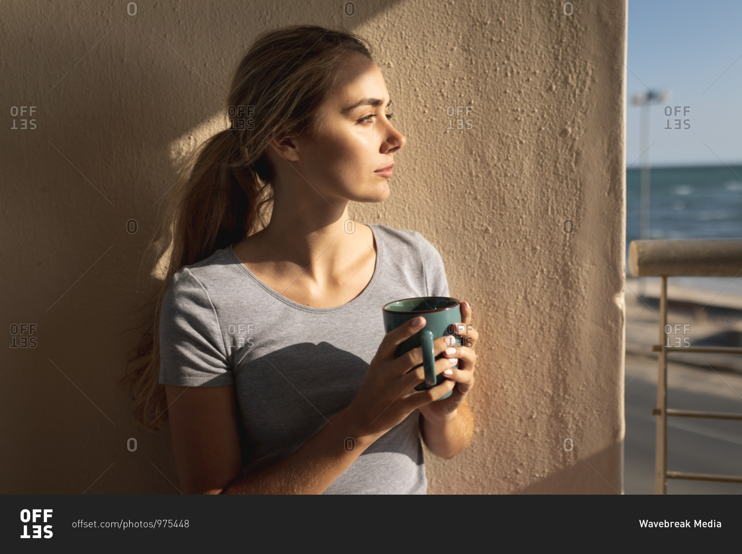 Caucasian woman standing on a balcony, holding a cup of coffee and looking away. Social distancing and self isolation in quarantine lockdown.