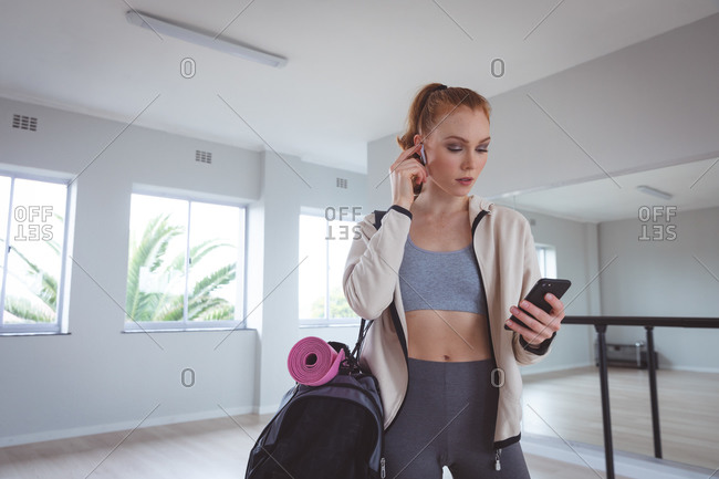 Caucasian attractive female ballet dancer with red hair wearing sportswear, entering a studio, preparing for a ballet class, looking at her phone with earphones in.