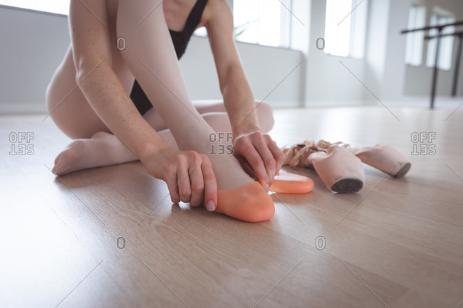 Caucasian attractive female ballet dancer putting on her ballet shoes, sitting on the floor, preparing for a ballet class in a bright studio.