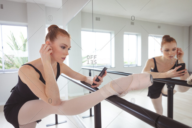 Caucasian attractive female ballet dancer with red hair holding a barre, preparing for a ballet class in a bright studio, stretching her leg and using her smartphone next to a mirror.