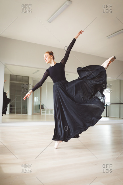 Caucasian attractive female ballet dancer with red hair dancing ballet, wearing a black, long dress, preparing for a ballet class in a bright studio, focusing on her exercise.