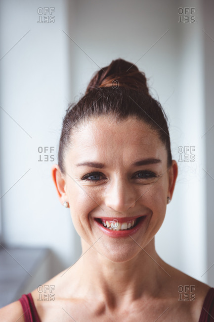 Portrait of a Caucasian attractive female ballet dancer with brown hair in a hair bun preparing for a ballet class in a bright studio, looking to camera with a smile on her face.
