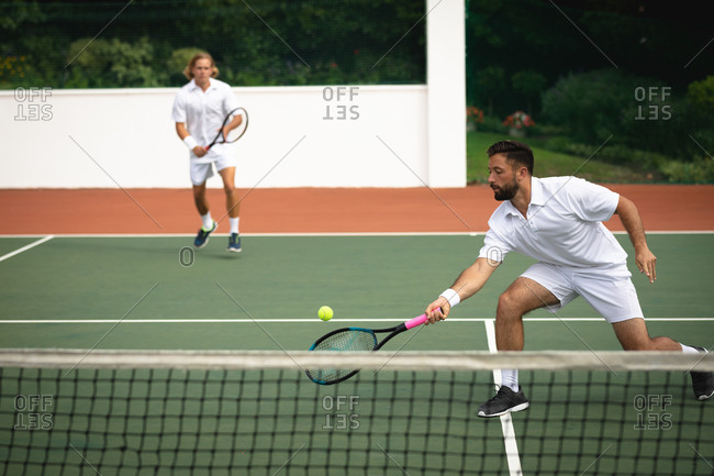A Caucasian and a mixed race men wearing tennis whites spending time on a court together, playing tennis on a sunny day, holding tennis, one of them hitting a ball