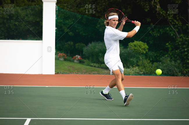 A Caucasian man wearing tennis whites spending time on a court playing tennis on a sunny day, holding a tennis racket and preparing to hit a ball