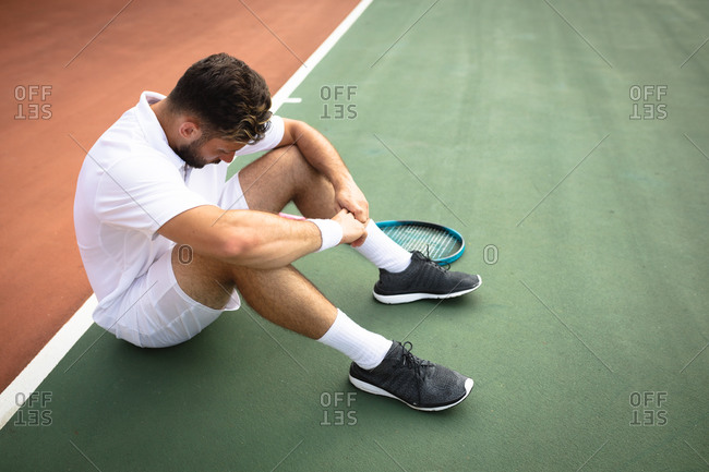 A mixed race man wearing tennis whites spending time on a court playing tennis on a sunny day, taking a break, sitting on a ground