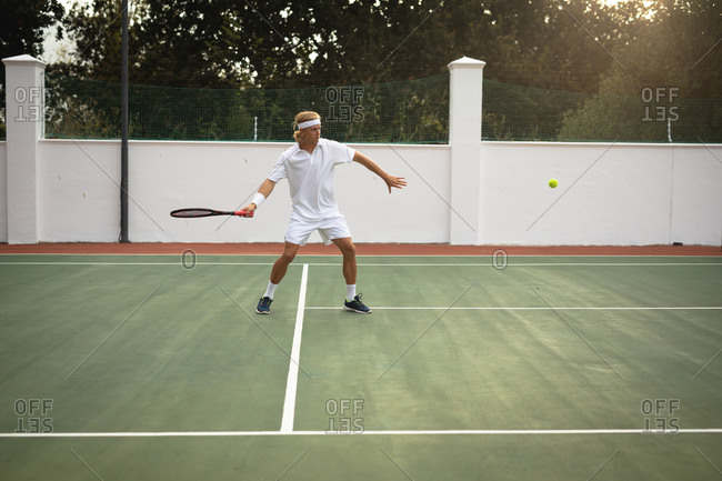 A Caucasian man wearing tennis whites spending time on a court playing tennis on a sunny day, holding a tennis racket, preparing to hit a ball