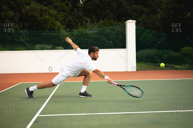 A mixed race man wearing tennis whites spending time on a court playing tennis on a sunny day, hitting a ball with a tennis racket