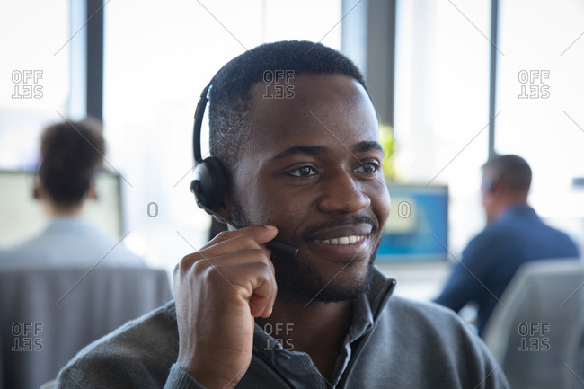 Close up of an African American businessman working in a modern office, sitting at a desk, wearing headset and talking, with his colleagues working in the background