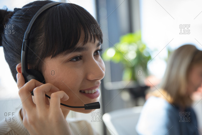 Close up of an Asian businesswoman working in a modern office, sitting at a desk, wearing headset and talking, with her colleague working in the background