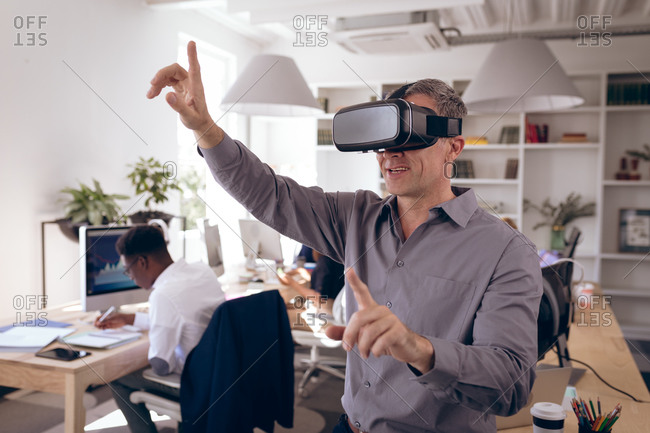 A Caucasian businessman working in a modern office, wearing VR headset, touching virtual interactive screen, with his business colleagues working at desks in the background