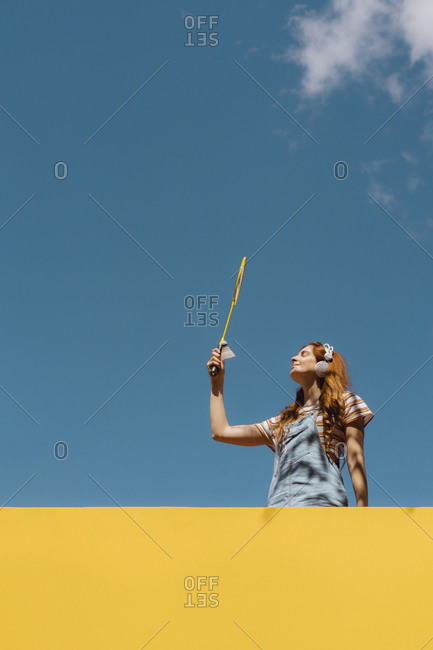 Young woman holding badminton racket while listening music above yellow wall against blue sky