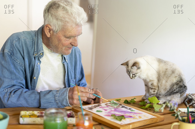 Senior man painting on paper while cat sitting on table at home
