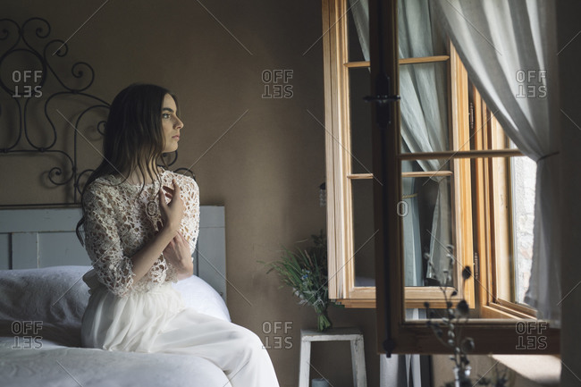 Young woman in elegant wedding dress sitting on bed looking out of the window
