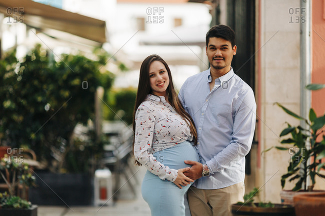 Happy expectant couple with hands on stomach standing in city during sunny day