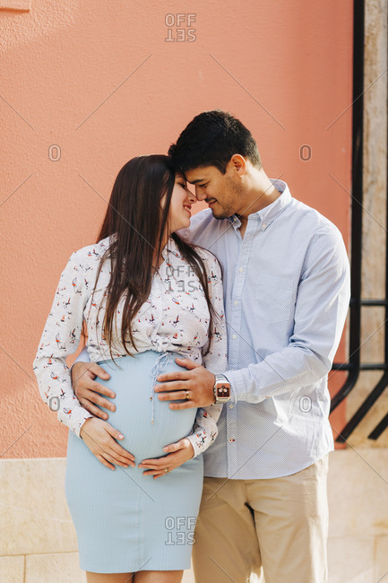 Romantic expectant couple standing with hands on stomach against pink wall in city