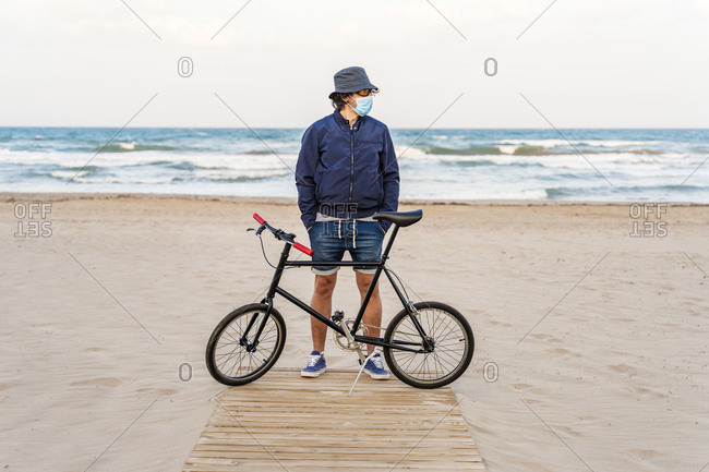 Mature man with bicycle- standing on the beach- wearing face mask