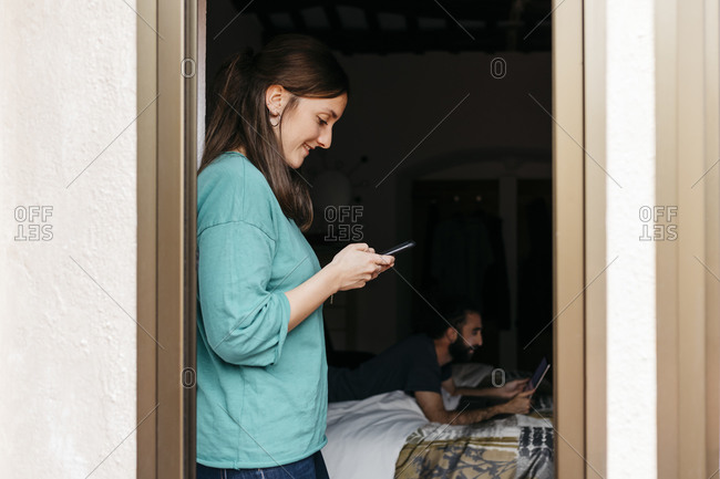 Woman using smartphone at open window- man using laptop lying on bed at home