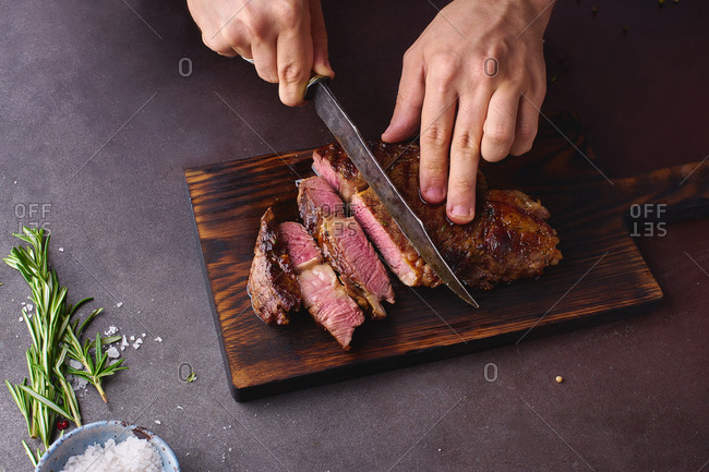 Male hands cutting cooked until medium ribeye steak on wooden cutting board.