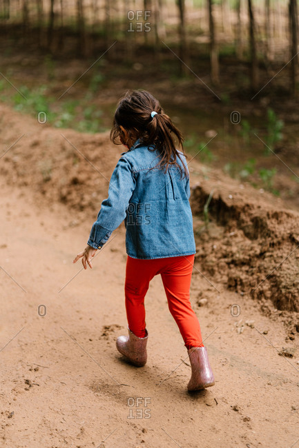 Back view of unrecognizable cute child in rubber boots strolling in mud path near dirty puddle