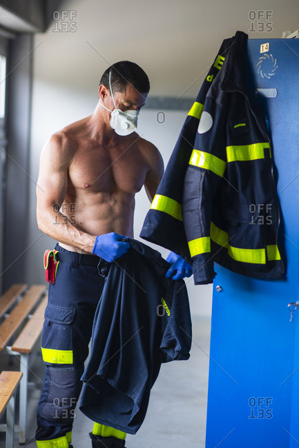 Serious fireman with naked torso and in latex gloves standing with respirator face mask near locker at fire station while preparing for work