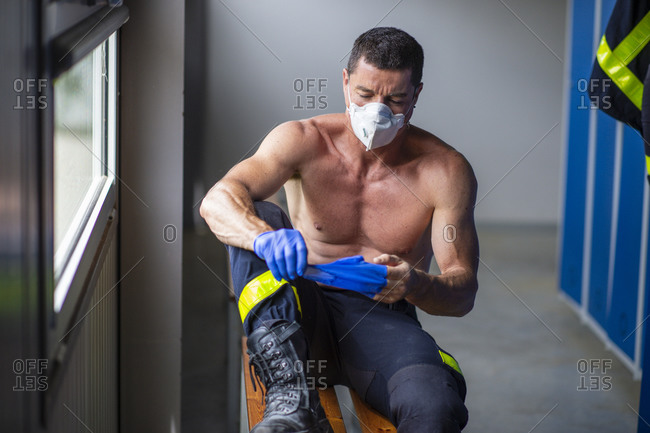 Serious fireman with muscular torso sitting on wooden bench at fire station and taking off latex gloves