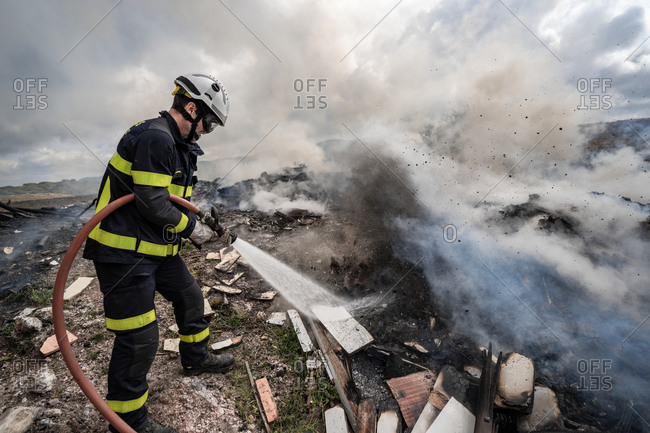 Side view of brave fireman in protective uniform standing with hose and extinguishing fire on dump in mountains