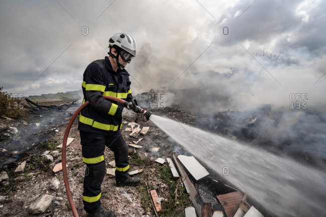 Side view of brave fireman in protective uniform standing with hose and extinguishing fire on dump in mountains