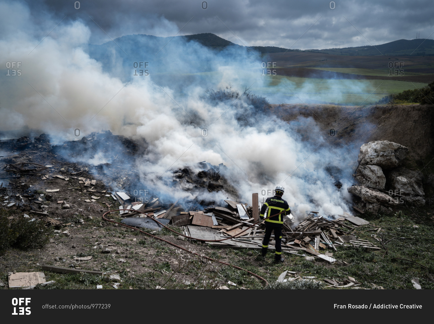 Back view of brave fireman in protective uniform standing with hose and extinguishing fire on dump in mountains