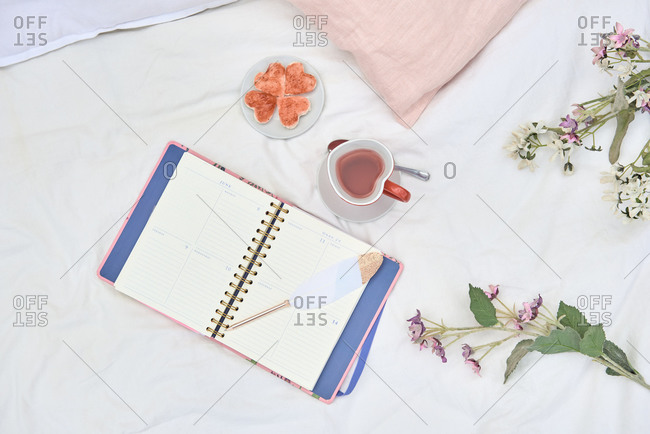 Top view of opened daily planner composed with cup of hot tea and tasty cookies placed on soft bed with flowers and cushion