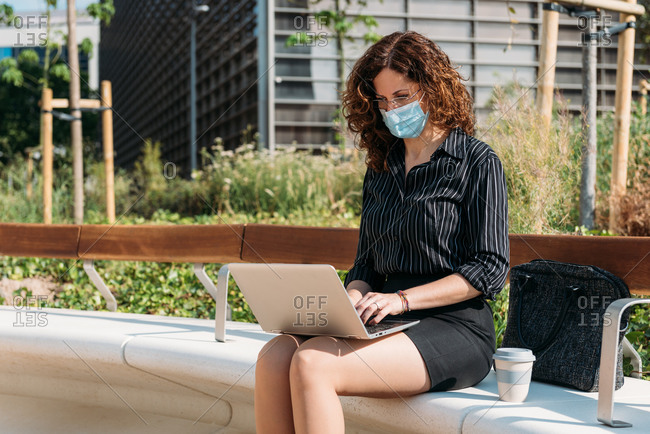 Business woman working on a park bench using her personal computer and wearing a protective mask.