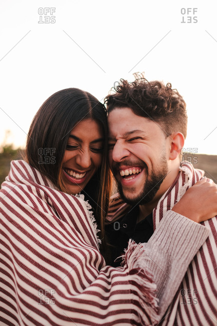 Delighted ethnic girlfriend and boyfriend embracing while covering with plaid and laughing at joke with closed eyes