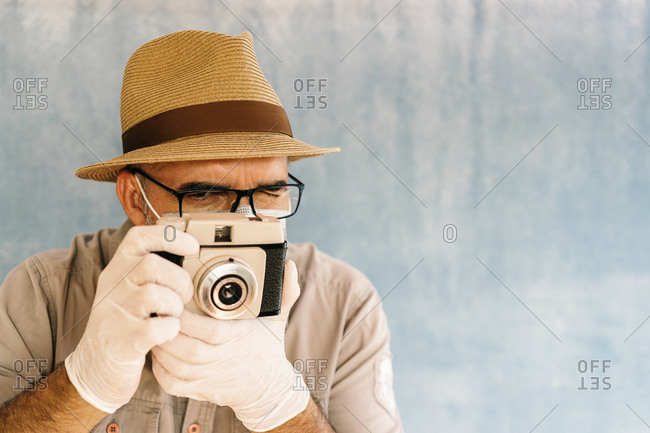 Middle aged man in latex gloves and medical mask taking picture with retro camera in light studio