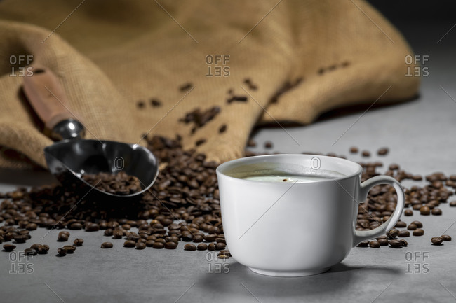 Fresh black coffee in white ceramic cup placed on saucer near coffee grinder and coffee beans on concrete table