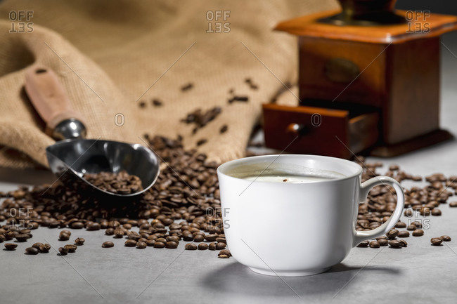Fresh black coffee in white ceramic cup placed on saucer near coffee grinder and coffee beans on concrete table