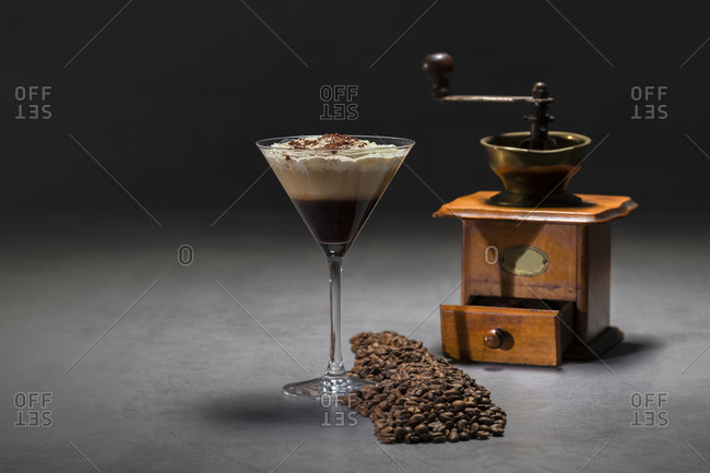 Roasted coffee beans and cocktail glass with yummy cappuccino with cream placed on concrete table near retro grinder