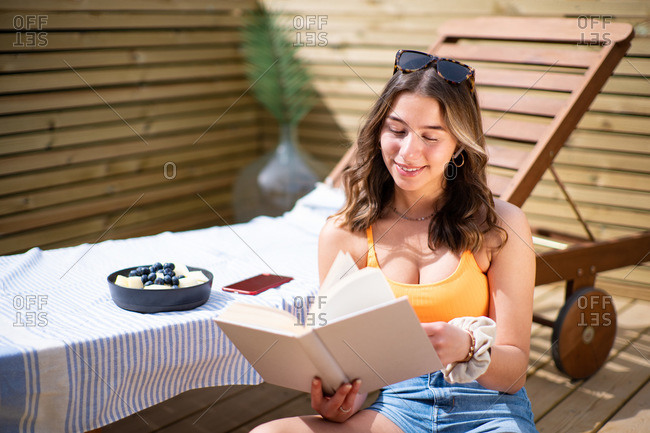 Cheerful female in summer top and shorts sitting near deck chair on wooden veranda and enjoying story while smiling and looking away