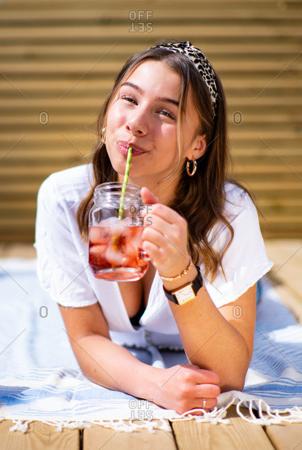 Cheerful young female in casual clothes drinking fresh fruit cocktail and enjoying sunny summer day while lying on terrace near instant photo camera