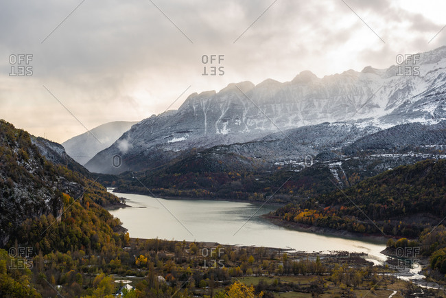 Lake and snowy mountain ridge with autumn tree located against overcast sundown sky in evening in nature