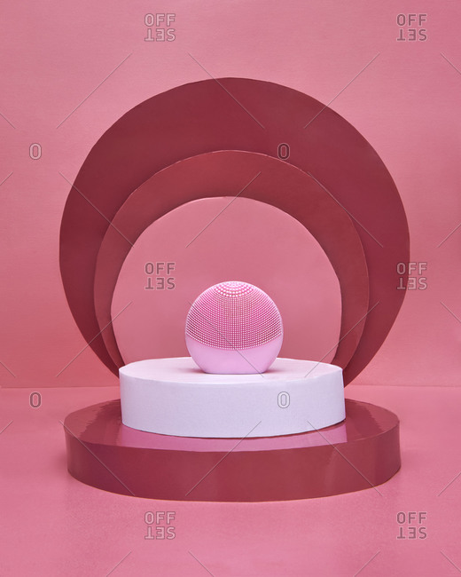 Pink electronic facial cleansing massager placed amidst composition with white and red disks on pink background