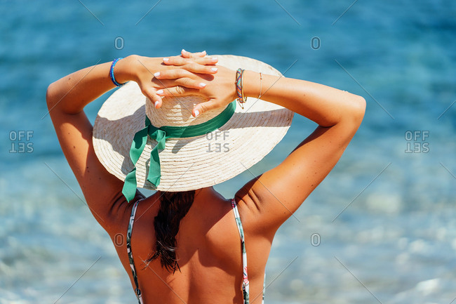 Back view of relaxed female tourist in straw hat and bikini standing on background of majestic seascape and admiring sea during summer holiday