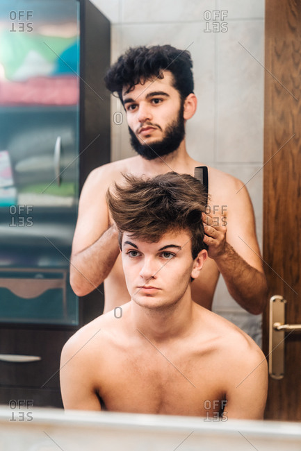 Man brushing hair of young guy and doing hairstyle in bright bathroom