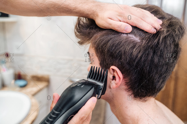 Male with hair trimmer cutting hair of guy in contemporary bathroom at home
