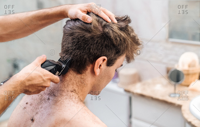 Male with hair trimmer cutting hair of guy in contemporary bathroom at home