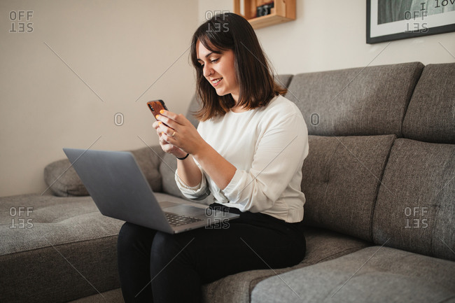 Confident female entrepreneur in casual clothes sitting on sofa in living room and using mobile phone while browsing on computer during remote work